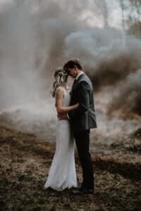 Ein Paar im Hochzeitskleid umarmt sich in einer nebligen Waldgegend. Die Braut trägt, während sie ihre Hochzeit sorgfältig plant, ein weißes Kleid, während der Bräutigam in einem dunklen Anzug neben ihr steht. Rauch oder Nebel schaffen einen dramatischen Hintergrund für diese bezaubernde Hochzeitsszene.