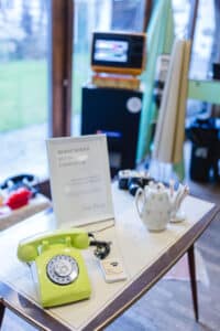 Ein Zimmer im Vintage-Stil, das sich perfekt für die Planung einer Hochzeit eignet. Auf dem Tisch steht ein grünes Telefon mit Wählscheibe neben einer gepunkteten Teekanne und Fotoapparaten. Spielkarten und ein gerahmtes Schild sorgen für Charme, während ein alter Fernseher neben dem Fenster steht und nostalgische Gefühle weckt, ideal für die Hochzeitsplanung.