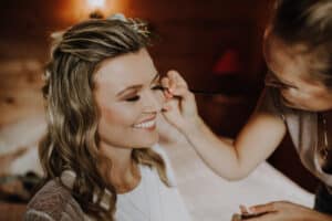 Eine Frau wird von einer Visagistin für ihre Hochzeit geschminkt. Sie lächelt mit geschlossenen Augen, während die Visagistin mit einem Pinsel sorgfältig Augen-Make-up aufträgt. Die Umgebung ist warm und gemütlich, mit sanftem Licht, perfekt für diesen besonderen Moment in ihrer Hochzeitsplanung.