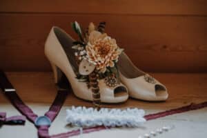 Cremefarbene Peep-Toe-Pumps, die mit einer großen Blume, Federn und Schmucksteinen geschmückt sind, stehen auf einer Holzoberfläche. Daneben befinden sich dunkelrote Hosenträger, ein Spitzenstrumpfband und ein paar kleine Verzierungen – perfekte Details für Ihr Hochzeits-Ensemble.