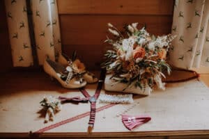 Eine wunderschön arrangierte Szene für die Hochzeit: Hochzeitsaccessoires auf einem Holztisch, darunter ein Strauß mit Rosen und getrockneten Blättern, beige, mit Blumen geschmückte High Heels, eine burgunderfarbene Fliege, Hosenträger, ein Strumpfband und eine Perlenkette.