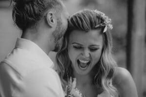 Ein Schwarzweißfoto zeigt ein Paar in einem freudigen Moment während der Hochzeit. Der Mann flüstert der Frau etwas ins Ohr und bringt sie zum Lachen. Sie hat Blumen im Haar, was zur unbeschwerten und intimen Atmosphäre dieses besonderen Tages beiträgt.