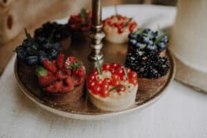 Auf einem mehrstöckigen Tablett für einen Hochzeitsempfang wird eine Auswahl kleiner Obsttörtchen präsentiert. Jedes Törtchen ist mit Himbeeren, Blaubeeren, Brombeeren, roten Johannisbeeren und weißen Johannisbeeren belegt, wodurch ein farbenfrohes und appetitanregendes Arrangement entsteht, das perfekt für die Feier ist.