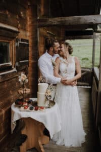 Braut und Bräutigam teilen einen zärtlichen Moment auf einer rustikalen Veranda und fangen damit die Essenz ihrer Hochzeit perfekt ein. Die Braut trägt ein Spitzenbrautkleid, während der Bräutigam ein weißes Hemd trägt. In der Nähe schmücken eine Torte und verschiedenes Gebäck einen Tisch, umgeben von Holzwänden und Grünpflanzen.