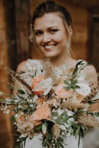 Eine Person hält einen Strauß verschiedener Blumen und Grünpflanzen und lächelt sanft. Der für eine Hochzeit entworfene Strauß enthält pfirsichfarbene Rosen, getrocknete Gräser und grüne Blätter, die ein rustikales und elegantes Arrangement ergeben. Der Hintergrund ist leicht verschwommen.