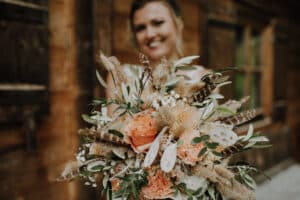 Eine lächelnde Frau hält einen rustikalen Blumenstrauß mit Trockenblumen, Federn und Grünzeug, perfekt für eine Hochzeit. Ein Holzgebäude bildet den Hintergrund und verleiht diesem Moment der Hochzeitsplanung Charme.