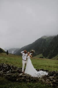 Ein Paar im Hochzeitskleid steht fröhlich auf einer Felswand in einem üppig grünen Tal, perfekt für ihre Hochzeitsplanung, mit nebelbedeckten Bergen. Die Braut hält einen Blumenstrauß und der Bräutigam trägt ein weißes Hemd und beige Hosen. Ein bewölkter Himmel verleiht ihrer Hochzeit eine dramatische Kulisse.