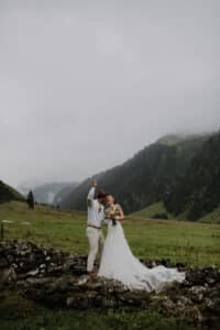 Ein Paar in Hochzeitskleidung steht auf einer Steinmauer in einem grünen Tal mit nebelverhangenen Bergen im Hintergrund. Während sie ihre Hochzeit feiern, hebt der Bräutigam in weißem Hemd und beiger Hose die Hand seiner Braut. Die Braut in einem fließenden Kleid hält einen Blumenstrauß, der die Romantik ihrer Hochzeit symbolisiert.