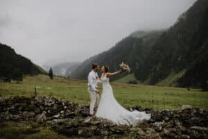 Ein Paar in Hochzeitskleidung steht auf einer Steinmauer in einem üppig grünen Tal, umgeben von nebligen Bergen. Die Braut, die ihren Blumenstrauß umklammert, gestikuliert freudig, während der Bräutigam sie liebevoll ansieht und damit die Essenz von Hochzeit und Feier verkörpert.
