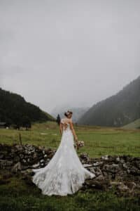 Eine Braut in einem weißen Spitzenhochzeitskleid steht mit ihrem Blumenstrauß in der Hand auf einer Wiese. Ihr Blick ist abgewandt, umgeben von nebligen Bergen und einem Steinzaun unter einem wolkigen Himmel und verkörpert die heitere Schönheit von Hochzeit inmitten der Umarmung der Natur.