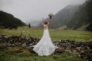 Eine Braut in einem weißen Spitzenbrautkleid steht mit dem Rücken zur Kamera, die Arme erhoben und einen Blumenstrauß in der Hand. Sie befindet sich in einer üppigen, bergigen Landschaft mit einer Steinmauer im Vordergrund und nebligen Hügeln, die die verträumte Kulisse für ihre Hochzeit bilden.