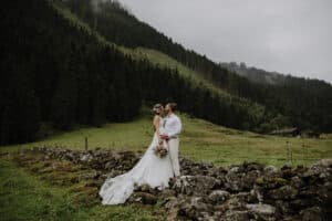 Braut und Bräutigam stehen auf einer Steinmauer in einer malerischen Berglandschaft und verkörpern ihre Hochzeitsträume. Das fließende weiße Kleid und der Blumenstrauß der Braut ergänzen den hellen Anzug des Bräutigams. Üppige grüne Hügel und ein Waldgebiet umgeben sie unter einem bewölkten Himmel, perfekt für ihre Hochzeitsplanung.