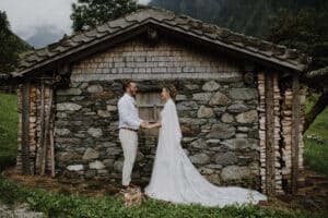 Ein Paar in Hochzeitskleidung hält sich vor einer rustikalen Steinhütte an den Händen und verkörpert damit die Essenz von „Heiraten“. Der Bräutigam trägt ein weißes Hemd und beige Hosen, während das weiße Kleid der Braut mit einer langen Schleppe die üppigen grünen Hügel ergänzt, die im Hintergrund wunderschön zu sehen sind.