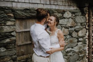 Ein Paar in Hochzeitskleidung umarmt sich und lächelt vor einer rustikalen Steinmauer mit einem Holzfenster und fängt die Essenz ihrer Hochzeit ein. Der Bräutigam trägt ein weißes Hemd und die Braut glänzt in einem weißen Spitzenkleid und hält einen kleinen Blumenstrauß in der Hand, was ihre sorgfältige Hochzeitsplanung verkörpert.