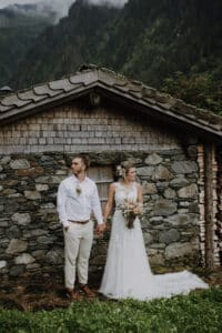 Braut und Bräutigam stehen Hand in Hand vor einer rustikalen Steinhütte. Die Braut hält einen Blumenstrauß und trägt ein weißes Kleid, während der Bräutigam ein weißes Hemd und helle Hosen trägt. In Anlehnung an ihre Traumhochzeit bilden üppige grüne Berge die perfekte Kulisse für ihre Liebesgeschichte.