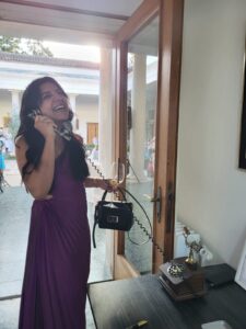 Eine Frau in einem lila Kleid steht lachend neben einer offenen Tür und hält ein antikes Telefon in der Hand. In ihrer anderen Hand balanciert sie eine kleine schwarze Handtasche und ein Getränk und genießt die Aufregung der Hochzeitsplanung. Im Hintergrund ist ein Gebäude mit Säulen und Grünflächen zu sehen, das die Bühne für ihre Hochzeit bildet.
