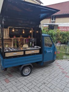 Ein kleiner blauer Kaffeewagen, perfekt für eine Hochzeit, steht auf einem gepflasterten Platz. Die Ladeklappe ist geöffnet und zeigt eine Theke mit einem Strohhut, Sirup und einer Kiste mit Lebensmitteln. Lichterketten hängen darüber und werfen ein warmes Licht vor dem Hintergrund eines Gebäudes mit rotem Dach.