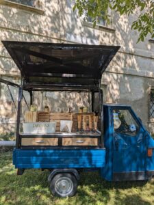Ein kleiner blauer Lastwagen steht auf dem Rasen und wurde in eine bezaubernde mobile Getränkestation für eine Hochzeit umgebaut. Er verfügt über Holzregale mit Bechern, Strohhalmen und einem „GETRÄNKE“-Schild. Im Schatten der Bäume neben einem alten Gebäude mit der Aufschrift „LOVE“ gelegen, ist er perfekt für alle, die heiraten möchten.