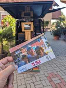 Eine Person hält eine Postkarte mit drei lächelnden Personen vor einem rustikalen Gebäude, möglicherweise aufgenommen während einer Hochzeit. Im Hintergrund sind ein offener Lastwagen, Topfpflanzen und ein gepflasterter Innenhof zu sehen. Auf der Postkarte steht elegant in Rot „95 Tage“.