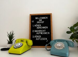 Zwischen einem grünen Wählscheibentelefon links und einem blauen Wählscheibentelefon rechts steht eine schwarze Buchstabentafel mit dem Text „WILLKOMMEN AM IRGHOF“ und einer Liste in deutscher Sprache. Passend für jede Hochzeit stehen Topfpflanzen neben den Telefonen auf einer weißen Fläche und schaffen eine einladende Atmosphäre.