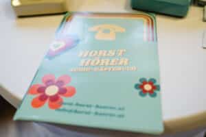 Eine Broschüre auf einem Tisch mit einem Vintage-Telefonsymbol sowie bunten Herz- und Blumenmotiven. Der Text lautet „HORST HÖRER AUDIO-GÄSTEBUCH“, perfekt für Ihre Hochzeitsplanung. Weitere Einzelheiten finden Sie im Website-Link und unter der E-Mail-Adresse unten.