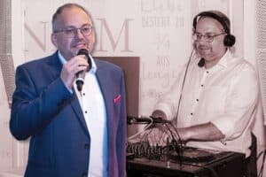 Ein Mann ist in zwei Rollen dargestellt: Links spricht er im Anzug bei einer Hochzeit in ein Mikrofon, rechts legt er mit Kopfhörern als DJ auf und orchestriert die Musik für die Hochzeitsplanung. Beide Szenen sind vor einem sanften rosa-beigen Hintergrund angesiedelt.
