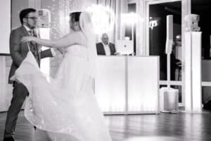 Ein Schwarzweißfoto zeigt ein Paar, das bei seiner Hochzeit tanzt. Die Braut in fließendem Kleid und Schleier und der Bräutigam in festlichem Anzug wirbeln anmutig in einem modernen Veranstaltungsort herum, während im Hintergrund ein DJ auflegt – ein Moment, der eine durchdachte Hochzeitsplanung widerspiegelt.