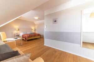 Ein gemütliches Schlafzimmer im Dachgeschoss mit Holzboden, perfekt für frisch Verheiratete, verfügt über ein Doppelbett mit einer rot-orange gemusterten Decke. Es umfasst zwei Nachttischlampen, ein beiges Sofa, einen kleinen Couchtisch und einen großen Spiegel an der rechten Wand, der das Licht vom Fenster reflektiert.