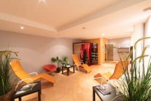 Ein entspannender Spa-Raum mit orangefarbenen Lounge-Sesseln, kleinen Tischen und Pflanzen ist ein idealer Ort zum Entspannen vor oder nach der Hochzeitsplanung. Eine Sauna im Hintergrund mit warmer Beleuchtung und beigefarbenem Fliesenboden verstärkt die sanfte Raumatmosphäre. Auf den Tischen liegen Zeitschriften.
