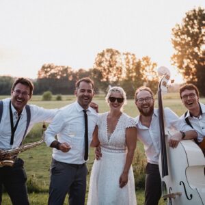 Eine Braut in einem weißen Kleid und mit Sonnenbrille posiert fröhlich mit vier Männern in Krawatte und Hosenträgern, die Musikinstrumente in der Hand halten, auf einer sonnigen Wiese – ein perfekter Moment, der Hochzeitsglück verkörpert. Bäume und ein strahlender Himmel dienen als bezaubernde Kulisse für diese idyllische Szene.