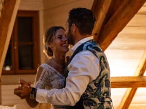 Ein Paar tanzt fröhlich in einem warm beleuchteten Raum mit Holzbalken und feiert seine Hochzeit. Die Frau trägt ein weißes Spitzenkleid, während der Mann ein Hemd und eine Weste mit Blumenmuster trägt. Beide lächeln und schaffen einen glücklichen und intimen Moment voller Liebe.