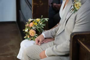 Ein Paar sitzt während einer Hochzeitszeremonie eng beieinander in einer Kirchenbank. Der Mann trägt einen hellgrauen Anzug mit einer Blumenboutonniere und die Frau hält einen bunten Blumenstrauß. Ihre Hände berühren sich sanft und symbolisieren Einheit und Liebe.