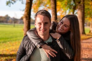 Ein Mann und eine Frau lächeln in einem sonnenbeschienenen Park und stellen sich ihre zukünftige Hochzeit vor. Die Frau sitzt spielerisch auf dem Mann, beide tragen Jacken. Im Hintergrund sind Gras, Bäume und Herbstblätter zu sehen, die eine warme, fröhliche Atmosphäre schaffen, die perfekt ist, um ihre Hochzeitsplanung zu besprechen.