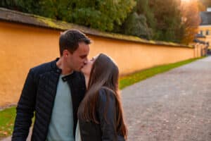 Ein Paar küsst sich romantisch auf einem von einer gelben Mauer und Bäumen gesäumten Weg im Freien, perfekt für ihre Hochzeitsfotos. Der Mann trägt eine schwarze Jacke, während sich die Frau in ihrer Lederjacke zu ihm beugt. Der Moment wird von warmem, sanftem Sonnenlicht erhellt.