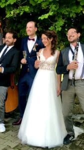 Eine Braut im weißen Kleid und ein Bräutigam im blauen Anzug mit Fliege lächeln und halten Champagnergläser in den Händen. Sie werden von zwei Musikern flankiert: einer mit einem Cello und der andere mit einer Klarinette, die eine zauberhafte Atmosphäre für ihre Hochzeit unter dem belaubten Baum schaffen.
