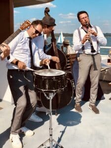 Eine Jazzband verleiht der Hochzeit im Freien eine lebendige Note, indem sie unter der Sonne spielt. Die Gruppe, bestehend aus einem Schlagzeuger, einem Kontrabassisten und einem Gitarristen mit Sonnenbrille, spielt neben einem See, an dem Segelboote vorbeigleiten – und verleiht der Hochzeitsfeier eine nautische Atmosphäre.