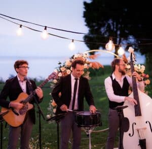 Eine dreiköpfige Band spielt im Freien unter Lichterketten, perfekt für jede Hochzeit. Die Musiker spielen Gitarre, Snare Drum und Kontrabass. Hinter ihnen befindet sich ein Blumenbogen mit malerischer Aussicht auf Wasser und Bäume. Sie sind in formelle Kleidung gekleidet und verleihen Ihrer Hochzeitsplanung einen eleganten Ton.