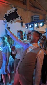Ein fröhlicher Mann in einem hellen Anzug jubelt mit erhobenem Arm und einem Stück schwarz-weißen Kuchen in der Hand. Um ihn herum tanzen die leger gekleideten Menschen und feiern eine scheinbar bezaubernde Hochzeit unter bunten Lichtern in einem rustikalen Holzsaal.