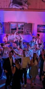 Eine lebhafte Hochzeitsfeier mit tanzenden und jubelnden Gästen. Im Hintergrund spielt eine Live-Band bei farbenfroher Beleuchtung. Der für Hochzeiten zugeschnittene Raum besticht durch rustikales Dekor, darunter eine Holzwand und Fenster mit Landschaftsfotos.