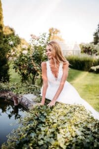 Eine Frau in einem weißen Brautkleid lehnt sich über einen kleinen Teich, der von üppigem Grün umgeben ist und verkörpert die Freude am Heiraten. Sie lächelt und genießt die sonnige Gartenkulisse, im Hintergrund sind Bäume und ein Gewächshaus zu sehen, und fängt einen perfekten Moment der Hochzeitsplanung ein.