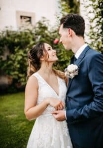 Eine Braut in einem Spitzenbrautkleid und ein Bräutigam in einem blauen Anzug stehen in einem Garten und fangen die Essenz ihrer Hochzeit ein. Während sie sich liebevoll anschauen, hält der Bräutigam einen kleinen Blumenstrauß inmitten grüner Büsche und Blumen in der Hand. Dies verkörpert Momente, die alle an der Hochzeitsplanung Beteiligten schätzen.