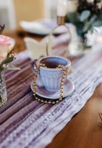 Eine zarte blaue Teetasse und Untertasse halten eine Kette aus goldenen Perlen auf einem Tisch, der mit einem violetten Tischläufer geschmückt ist, perfekt für eine elegante Hochzeit. Daneben stehen sanft akzentuierte Blumen, die dem Hochzeitsdekor Charme verleihen.