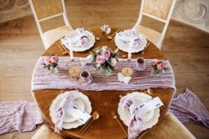 Ein runder Holztisch für vier Personen mit hellvioletten Tischdecken und Blumenarrangements ist perfekt für eine Hochzeit. Jedes Gedeck umfasst einen weißen Teller, ein goldfarbenes Besteck und eine violette Serviette. Rosa und violette Rosen in kleinen Vasen schmücken die Mitte, ideal für eine exquisite Hochzeitsplanung.