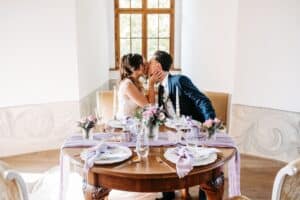 Braut und Bräutigam küssen sich während ihrer Hochzeit an einem kleinen, elegant dekorierten runden Tisch. Der Tisch ist mit rosa und lila Blumen, eleganten Gedecken und einem lavendelfarbenen Tischläufer geschmückt. Sonnenlicht fällt durch ein Fenster hinter ihnen herein.