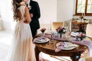 Braut und Bräutigam küssen sich neben einem elegant für ihre Hochzeit gedeckten Tisch, geschmückt mit Blumenarrangements, Tellern und lavendelfarbenen Servietten in einem sonnendurchfluteten Raum. Das Kleid der Braut ist mit Spitzendetails versehen, während sanftes Licht durch die Fenster fällt.
