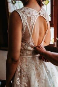 Eine Person, die ein Spitzenbrautkleid mit offenem Rücken trägt, wird beim Zuknöpfen unterstützt – perfekt für eine Traumhochzeit. Das Kleid ist mit aufwendigen Blumenmustern verziert und Buntglasfenster sorgen für eine warme Kulisse.