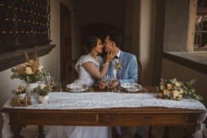 In einem warm beleuchteten, mit Blumenarrangements geschmückten Raum sitzt ein Paar an einem für das Essen gedeckten Holztisch. Die Frau in ihrem weißen Kleid und der Mann in seinem blauen Anzug sind formell gekleidet und blicken sich liebevoll in die Augen, was die Essenz ihrer Hochzeitsfeier einfängt.