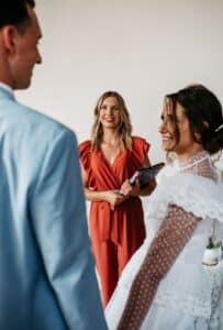 Ein Paar im Hochzeitskleid steht einander gegenüber, während eine Frau in einem roten Kleid sie anlächelt und möglicherweise die Zeremonie leitet. Die Braut trägt ein weißes Kleid und der Bräutigam einen hellblauen Anzug – perfekte Elemente für ihre wunderschöne Hochzeit.