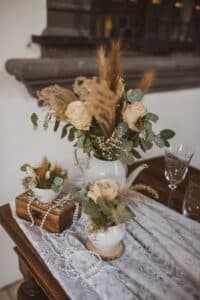 Blumenarrangements in weißen Keramikkrügen auf einem Holztisch mit Spitzendecke geben den perfekten Ton für eine romantische Hochzeit an. Die Sträuße aus Rosen und getrockneten Gräsern zusammen mit einer Holzkiste, Perlensträngen und Kristallgläsern schaffen eine bezaubernde Szene, die sich ideal für eine Hochzeit eignet.