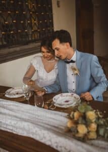 Braut und Bräutigam sitzen zusammen an einem wunderschön dekorierten Tisch und sind vertieft in ihre Hochzeitsplanung, während sie das Menü durchsehen. Die Braut glänzt in einem weißen Spitzenkleid, während der Bräutigam in einem hellblauen Anzug und einer Fliege elegant aussieht. Elegantes Geschirr und ein Blumenschmuck runden die intime Hochzeitsatmosphäre ab.